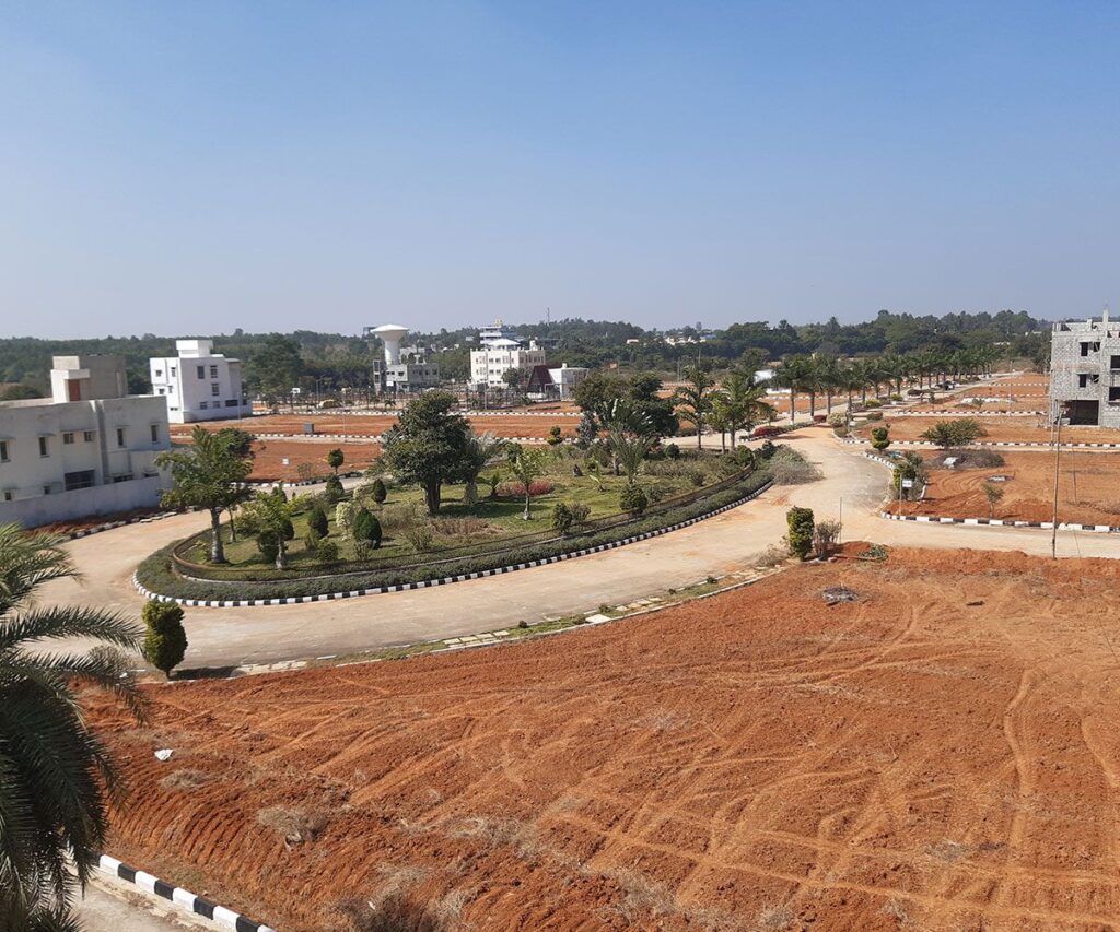 esidential plotted development viewed from above, featuring landscaped roads, landscaped roundabout, open plots, streetlights, palm trees, and surrounding low-rise buildings.